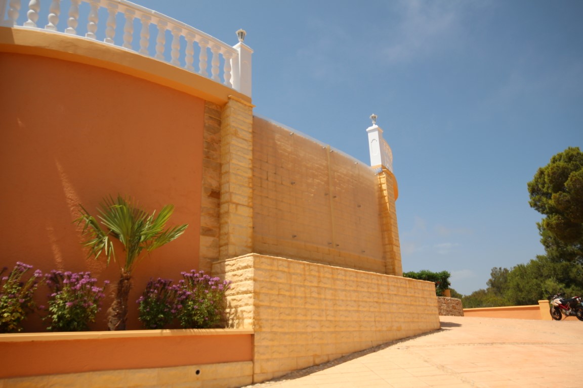 Vista exterior con muro de piedra tosca, pantalla protectora de cristal y balaustrada blanca, complementada por una jardinera con palmera y flores.