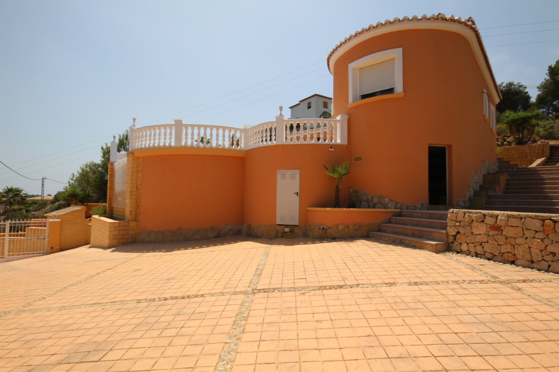 Vista exterior de la villa con fachada color terracota, arquitectura curva y amplio patio pavimentado. Incluye muros de piedra natural y balaustradas blancas en la terraza superior.