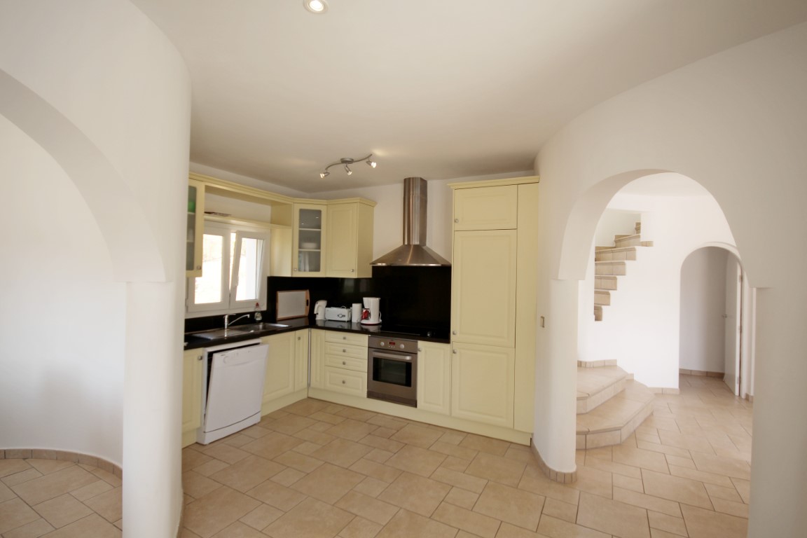 Cocina equipada con armarios color crema, encimera oscura y electrodomésticos integrados. El espacio destaca por sus arcos arquitectónicos y acceso directo a la zona de escaleras.