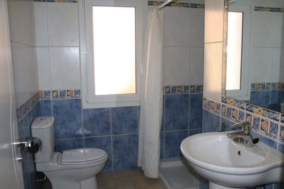 Cuarto de baño con azulejos azules y blancos, plato de ducha con cortina, lavabo de pedestal y ventana de cristal translúcido que aporta luz natural.