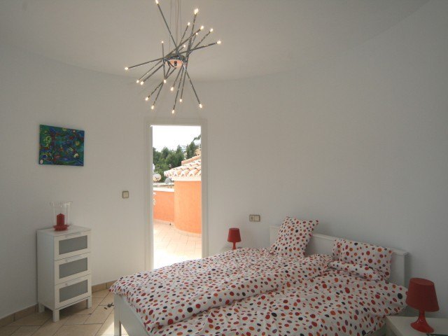 Dormitorio luminoso con paredes curvas, lámpara de diseño moderno y acceso directo a una terraza privada. Incluye mobiliario blanco y suelos cerámicos.
