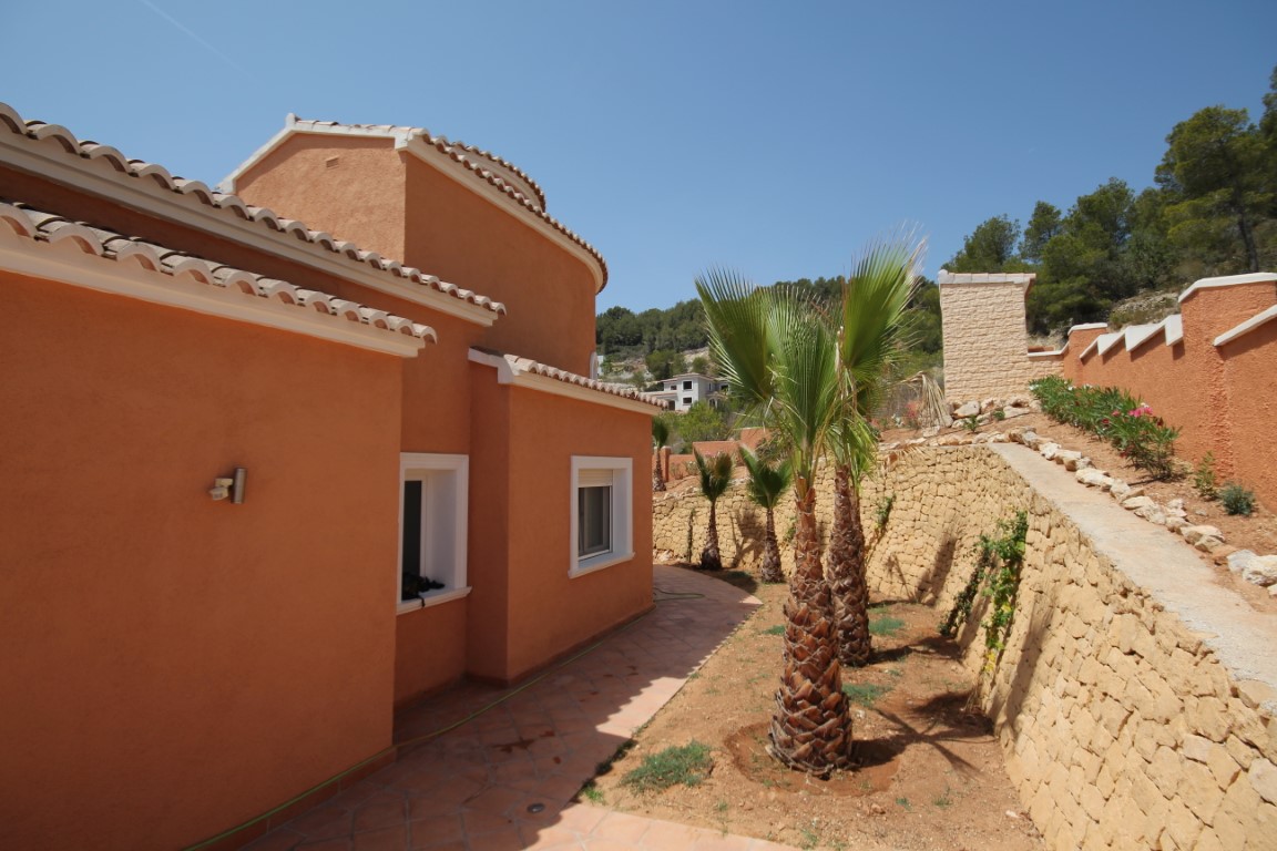 Fachada de color terracota con tejados de teja cerámica, acompañada de un jardín con palmeras y muros de piedra natural en varios niveles.