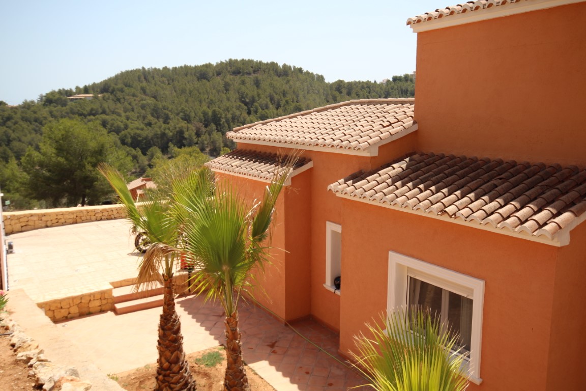 Fachada de estilo mediterráneo en tonos terracota con tejados de teja cerámica, situada junto a un patio pavimentado con palmeras y vistas a las colinas boscosas.