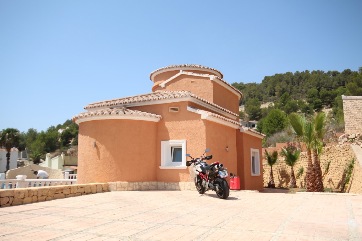 Fachada exterior de la villa con paredes curvas en tono terracota, zona de aparcamiento pavimentada y muros de piedra natural bajo un cielo despejado.