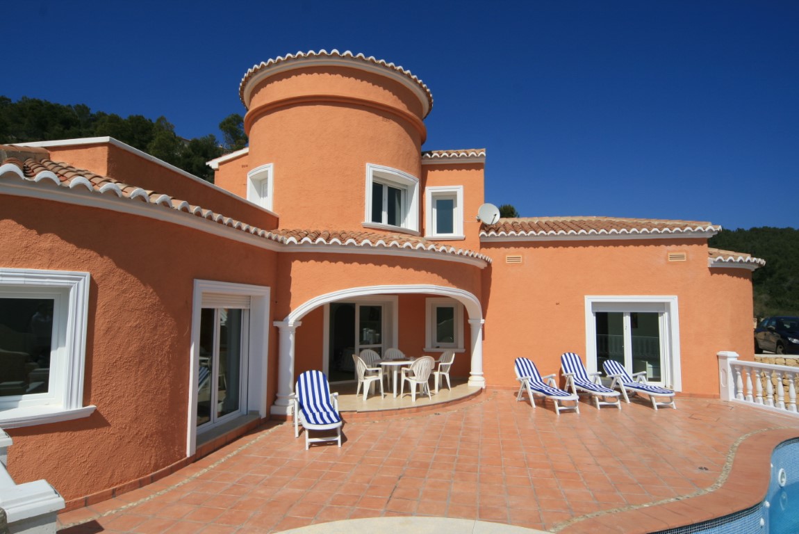 Fachada mediterránea en tonos terracota con torre circular, amplia terraza de baldosas cerámicas, zona de comedor bajo arco y tumbonas junto a la piscina.