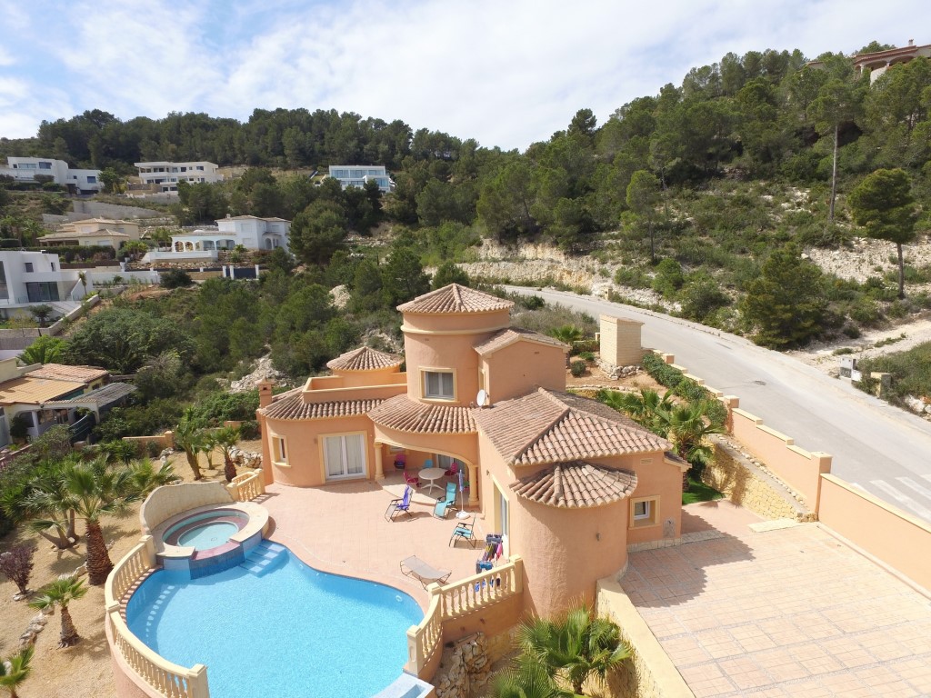 Vista aérea de la villa de estilo mediterráneo con piscina curva, jacuzzi integrado y amplia terraza solárium en una parcela elevada rodeada de pinos.