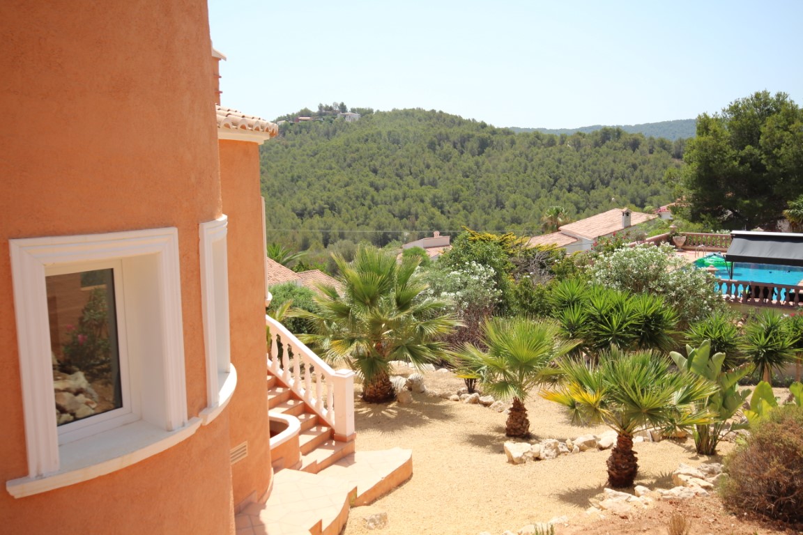 Vista exterior de la fachada curva en tonos terracota con jardín mediterráneo de bajo mantenimiento, palmeras y vistas despejadas al valle boscoso.