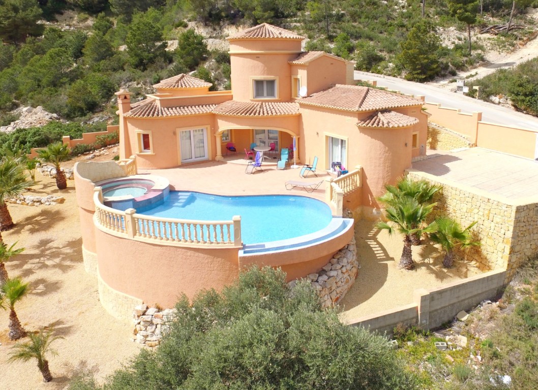 Vista exterior de la villa con fachada mediterránea, destacando la piscina infinita curva con jacuzzi integrado y amplia terraza solárium.