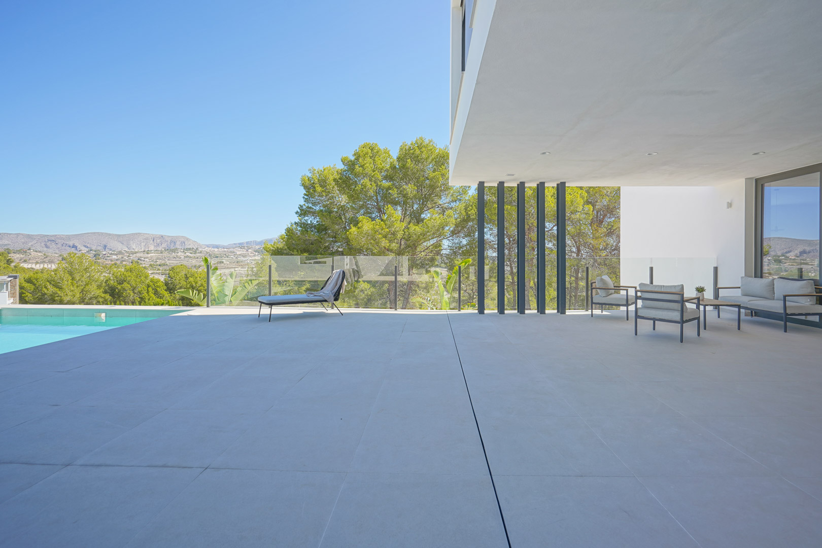Amplia terraza con suelo de piedra, zona de estar cubierta con mobiliario moderno y vistas despejadas a la pinada mediterránea junto a la piscina.
