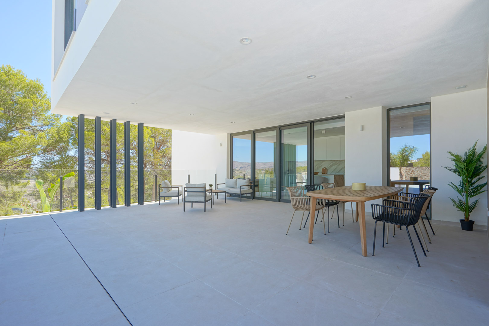 Amplia terraza cubierta con suelos cerámicos, zona de comedor exterior y salón lounge, protegida por barandillas de cristal y rodeada de pinos.