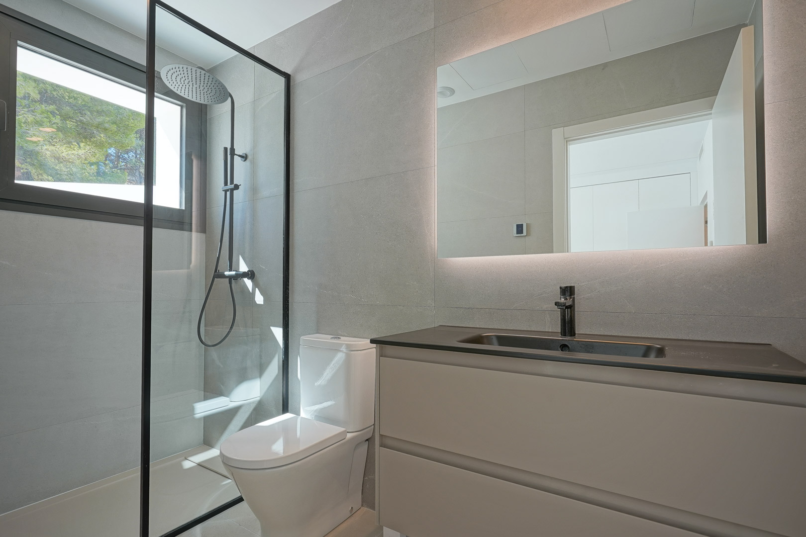 Baño moderno con azulejos grises, ducha de obra con perfilería negra y espejo retroiluminado. Incluye lavabo con encimera oscura y mueble suspendido en tonos neutros.