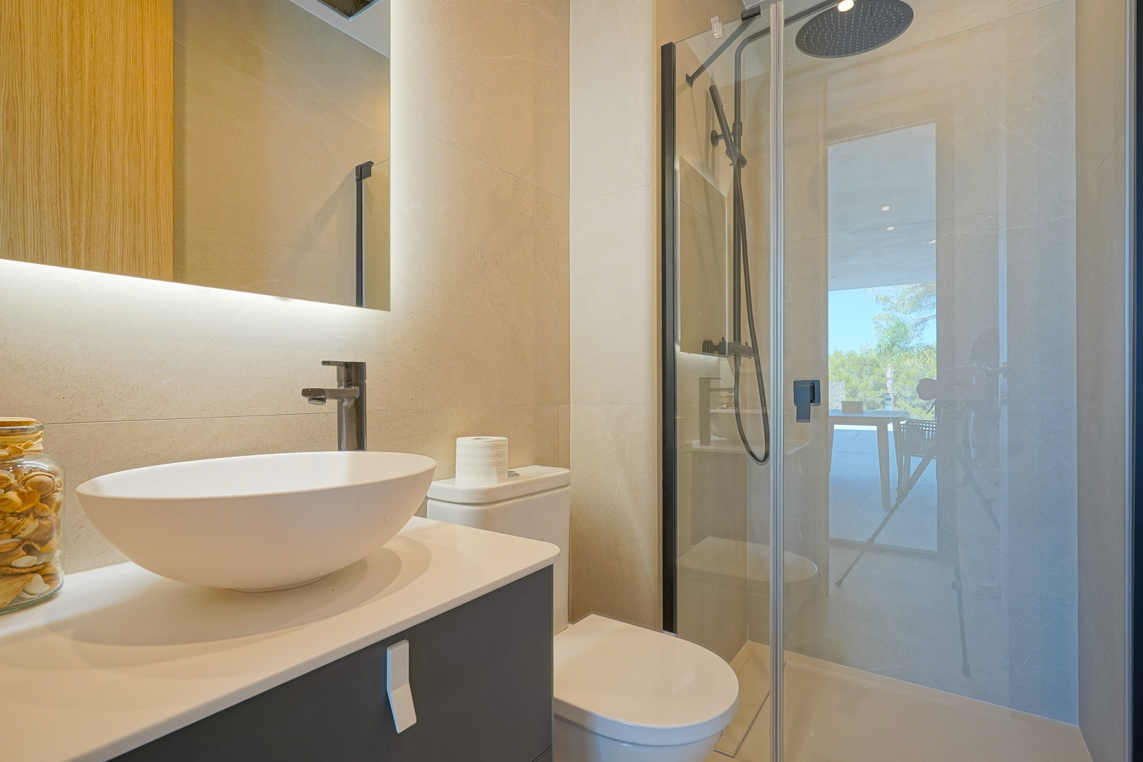 Baño moderno con lavabo sobre encimera, espejo retroiluminado y ducha con mampara de cristal. Los tonos crema y la grifería negra aportan un diseño elegante y funcional.