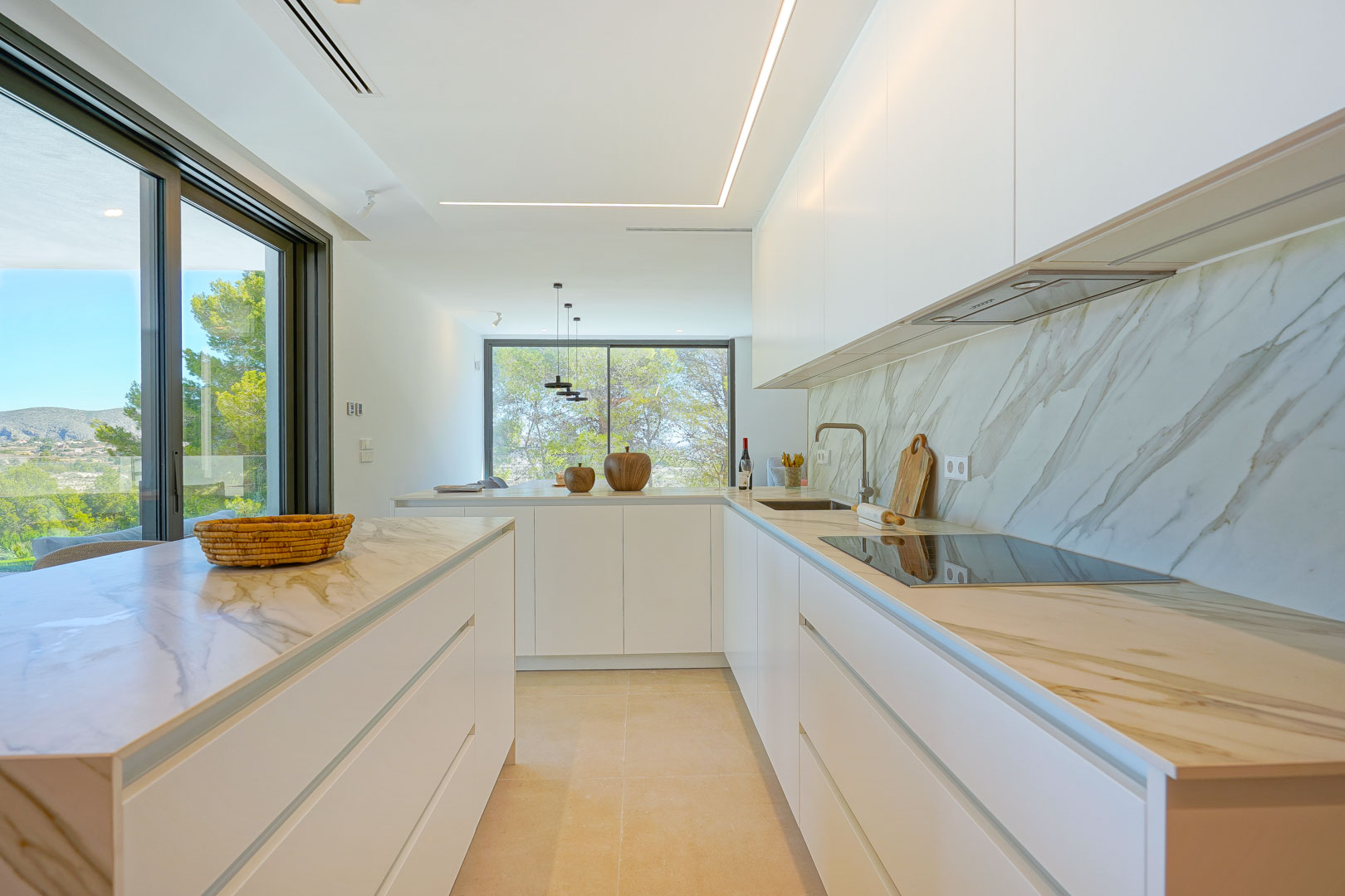 Cocina americana minimalista con encimeras de efecto mármol, armarios blancos sin tiradores e iluminación LED lineal. Grandes ventanales ofrecen vistas al entorno boscoso.