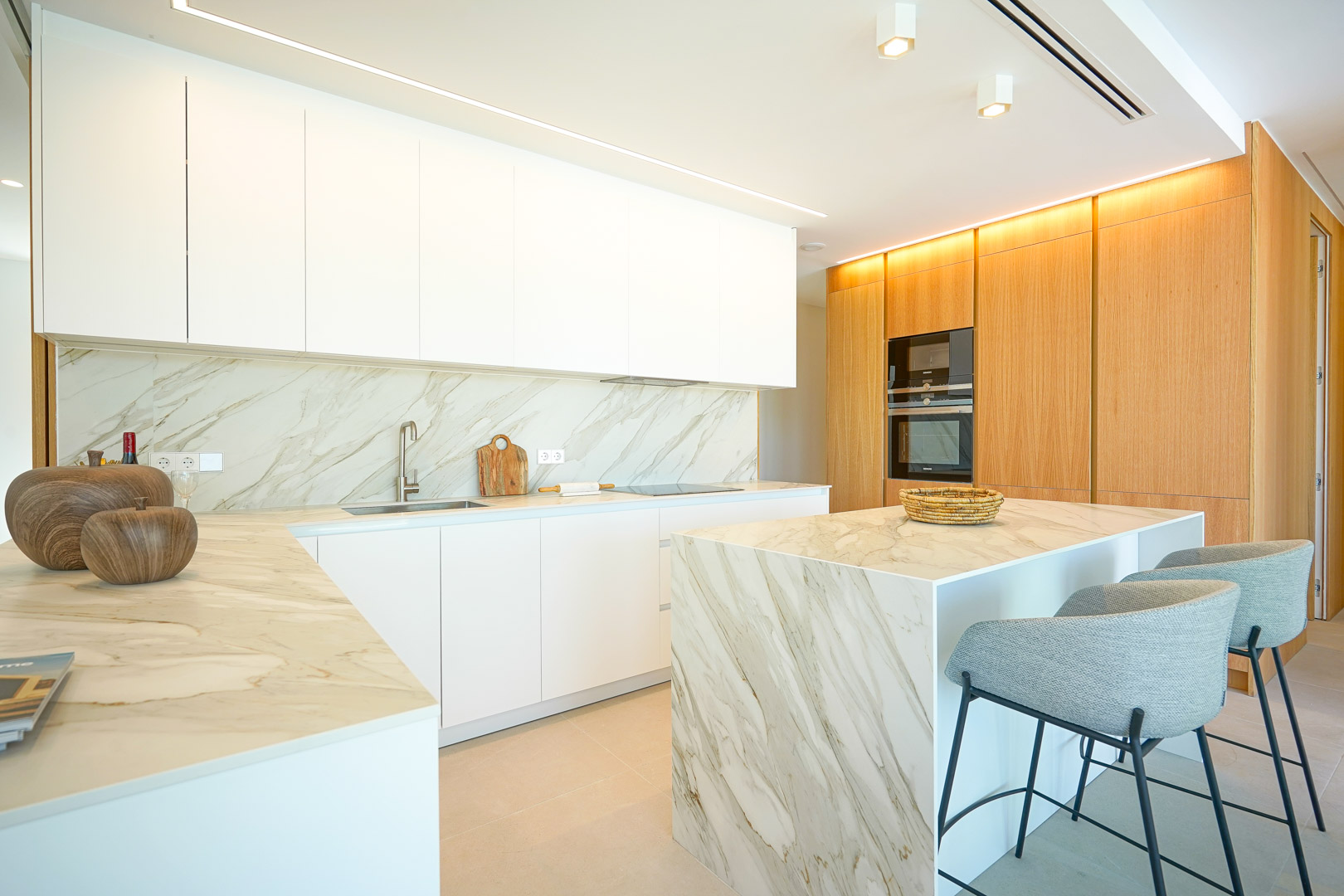 Cocina moderna con armarios blancos sin tiradores, encimeras de efecto mármol y una isla a juego. Un panelado de madera integra electrodomésticos bajo iluminación LED indirecta.