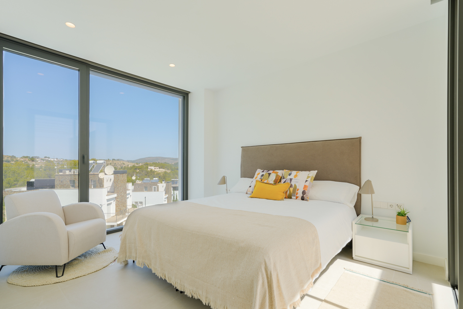 Dormitorio luminoso con ventanal de suelo a techo, cama doble con cabecero tapizado y sillón de lectura. Ofrece vistas despejadas al entorno natural de Moraira.