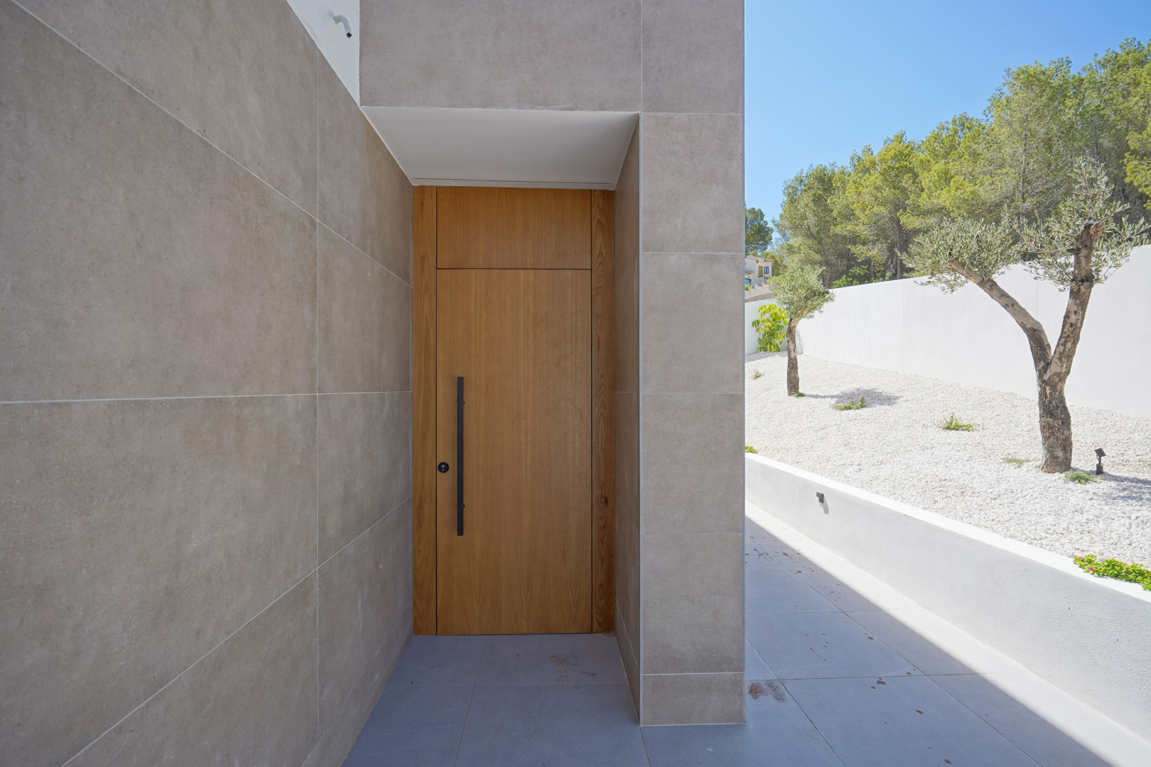 Entrada principal con puerta de madera maciza y tirador negro, enmarcada por fachada de gres porcelánico y jardín de grava blanca con olivos.