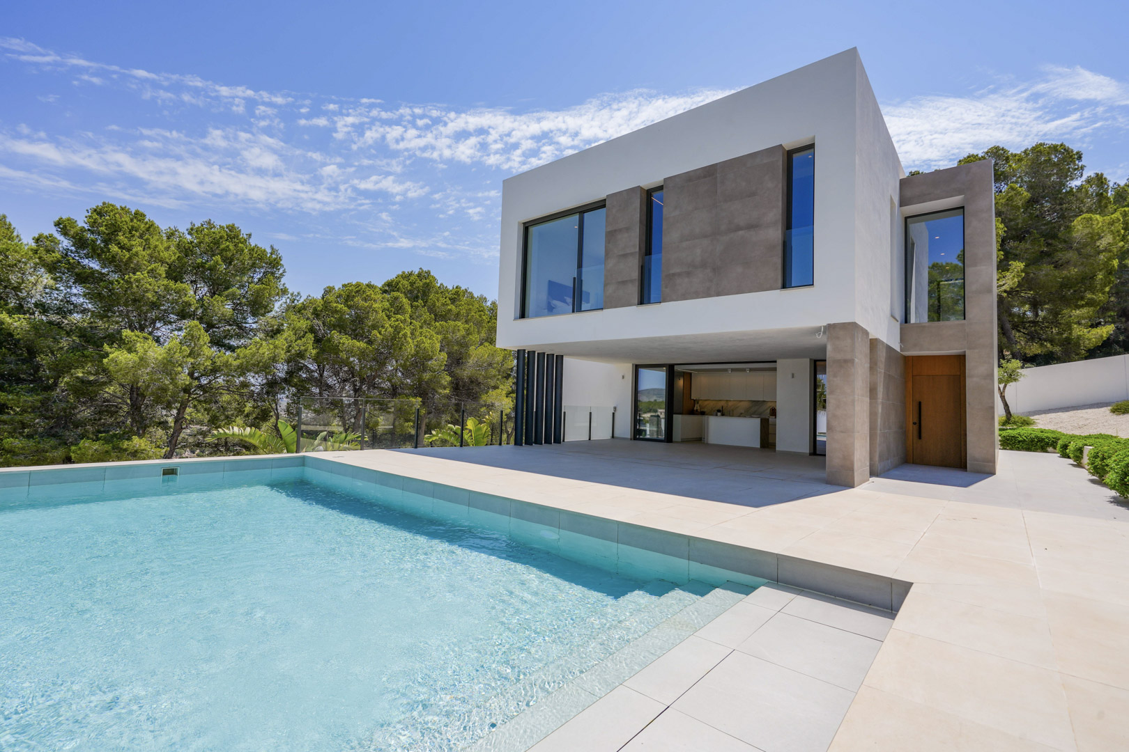 Exterior de la villa con piscina de borde infinito y amplia terraza de piedra clara, destacando su arquitectura moderna rodeada de un frondoso pinar.