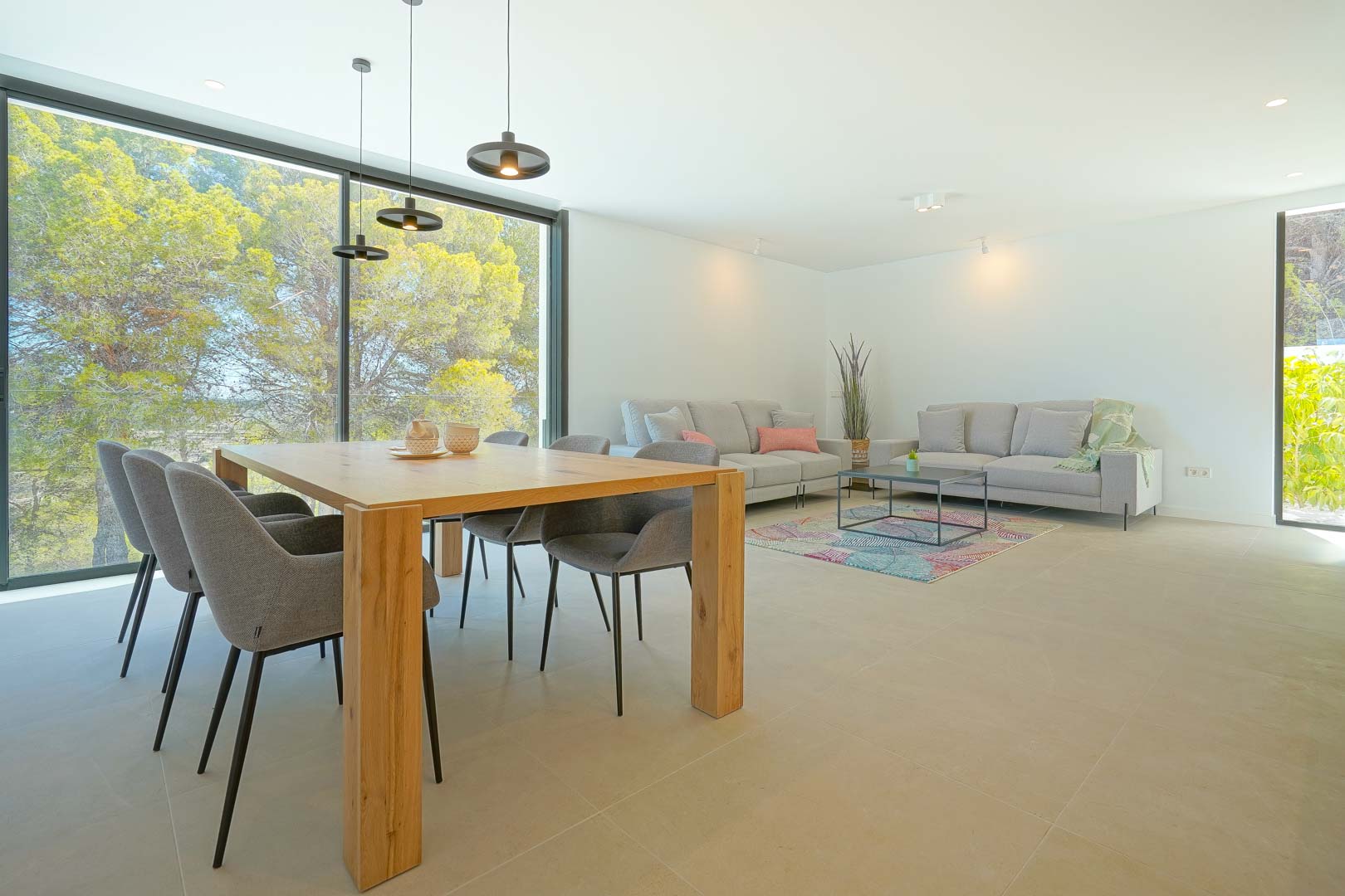 Salón-comedor diáfano con mesa de madera, sillas tapizadas y sofás grises. Grandes ventanales integran el entorno boscoso con el interior minimalista de la villa.