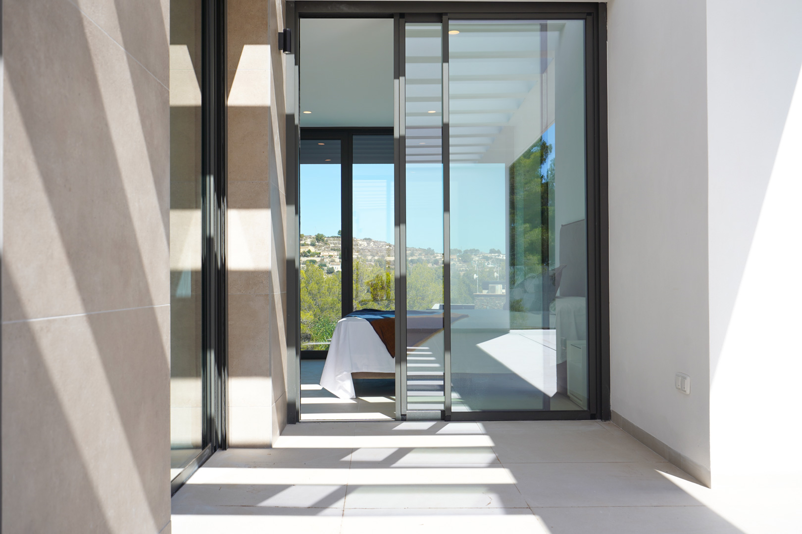 Terraza con fachada de piedra y puertas correderas de cristal que dan acceso a un dormitorio. Sombras lineales de la pérgola sobre el suelo cerámico.