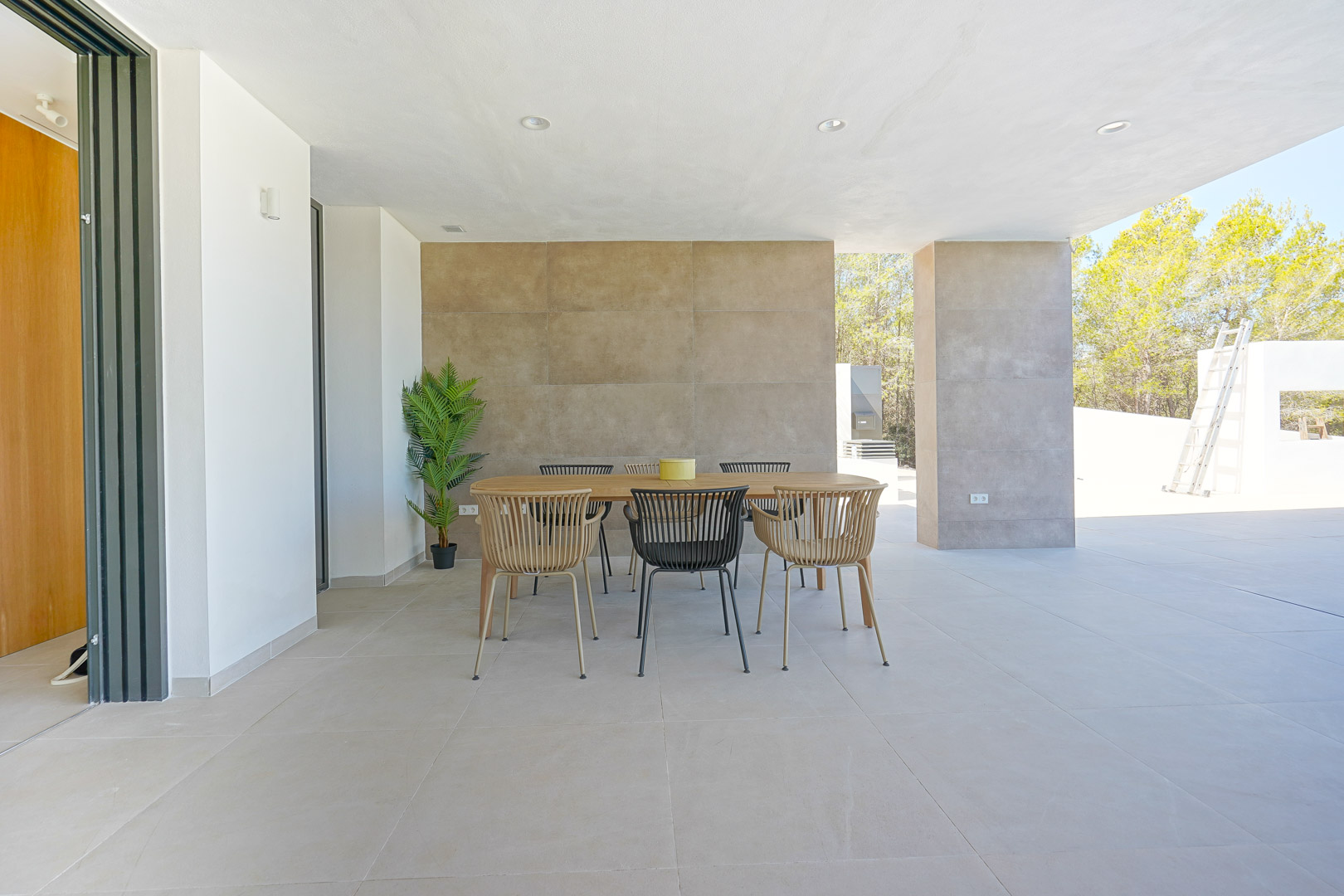 Terraza cubierta con zona de comedor exterior, pavimentada con gres porcelánico y pared decorativa texturizada. Iluminación integrada y vistas al entorno boscoso.