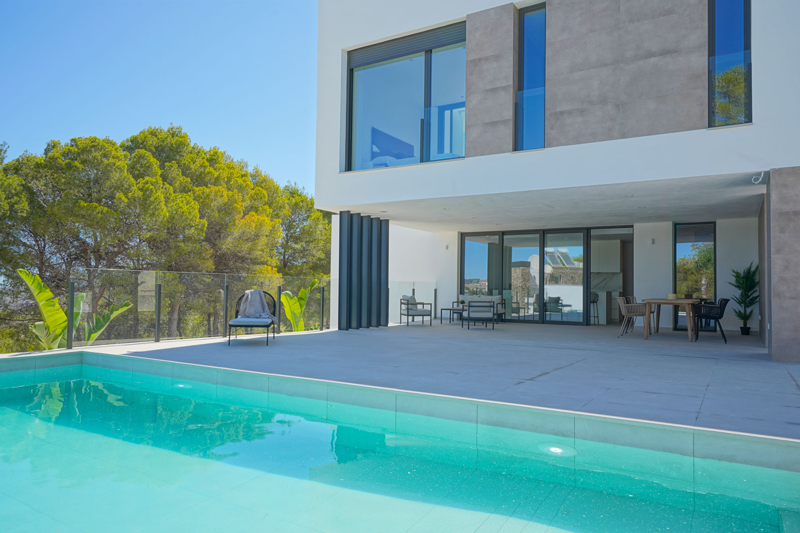 Terraza principal junto a la piscina con zona de estar y comedor exterior, bajo una fachada moderna de líneas rectas y rodeada de vegetación mediterránea.