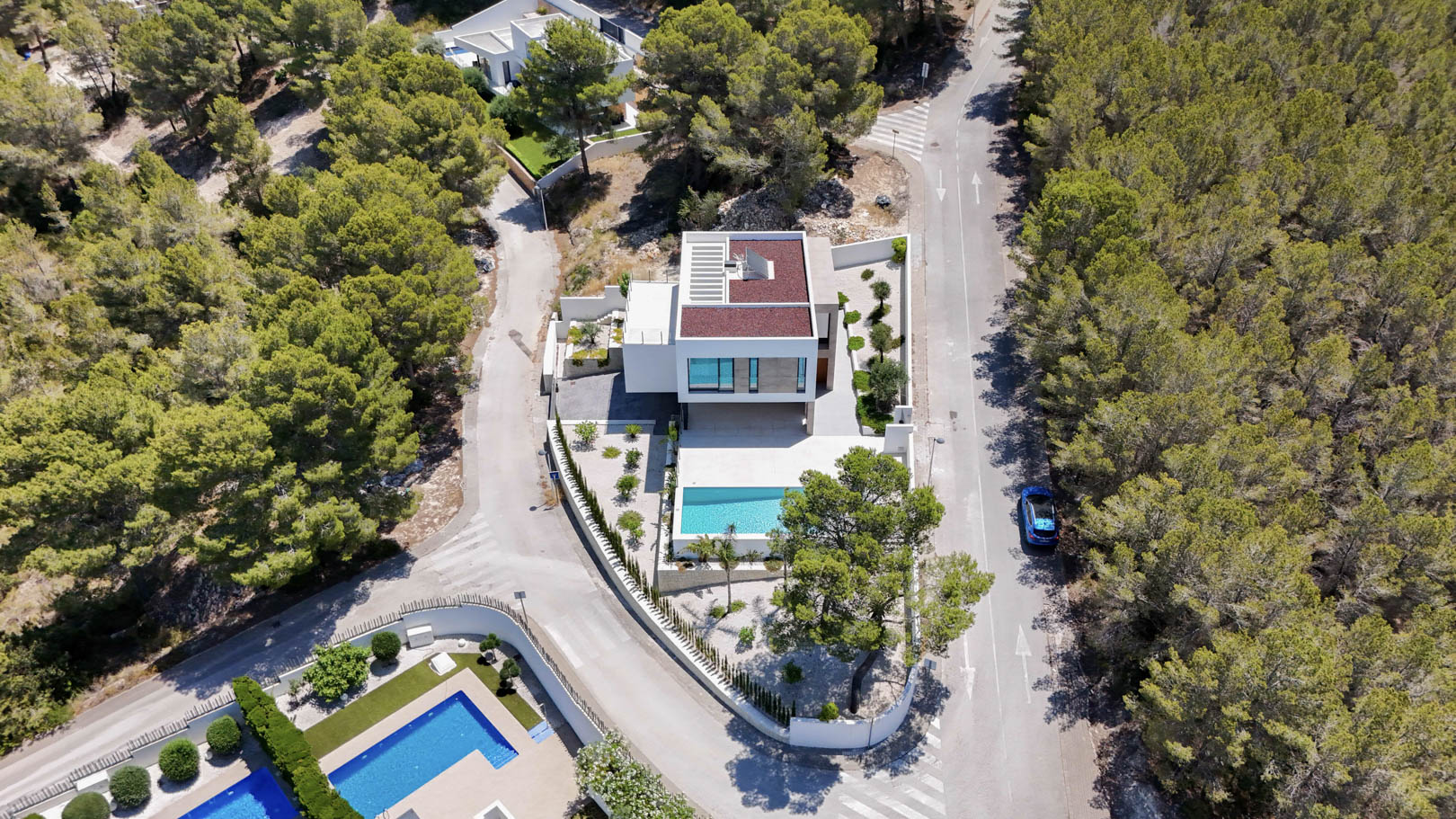 Vista aérea de la villa moderna situada en una parcela de esquina rodeada de pinos, con piscina rectangular, terrazas blancas y jardín mediterráneo.