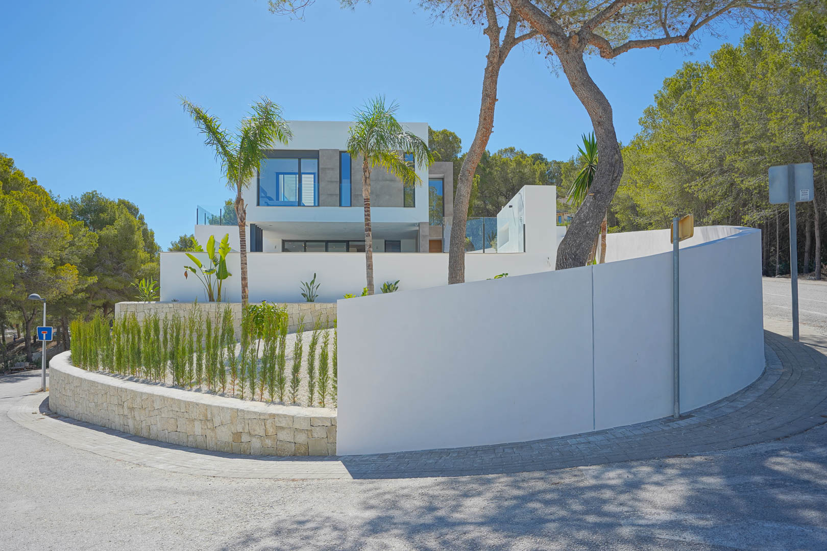 Vista exterior de la villa con fachada blanca, muros perimetrales curvos y jardín mediterráneo con cipreses y palmeras en un entorno boscoso.