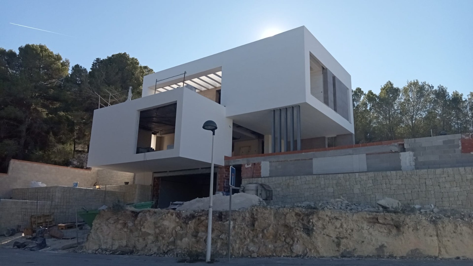 Vista exterior de la villa en construcción, destacando su arquitectura cubista con volúmenes en voladizo sobre un muro de piedra natural y garaje.