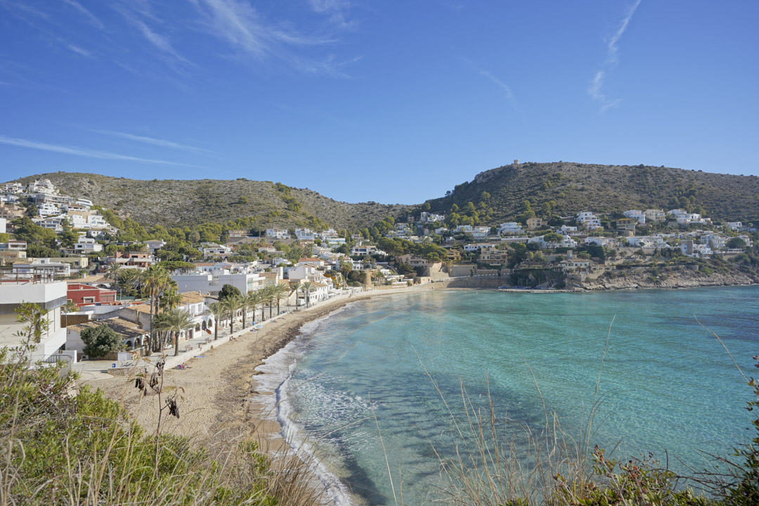Vista panorámica de la playa de El Portet en Moraira, destacando sus aguas cristalinas, el paseo marítimo con palmeras y las colinas residenciales circundantes.