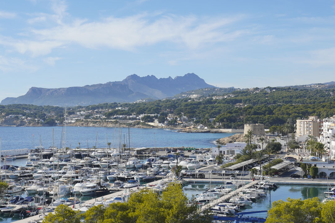 Vistas panorámicas del puerto deportivo de Moraira y el Castillo, con la Sierra de Bernia al fondo bajo un cielo despejado.