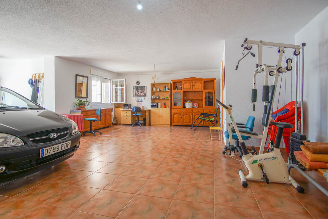 Amplio espacio multifuncional en la planta baja con suelos cerámicos, que integra zona de garaje, área de gimnasio y mobiliario de madera maciza para oficina o almacenaje.