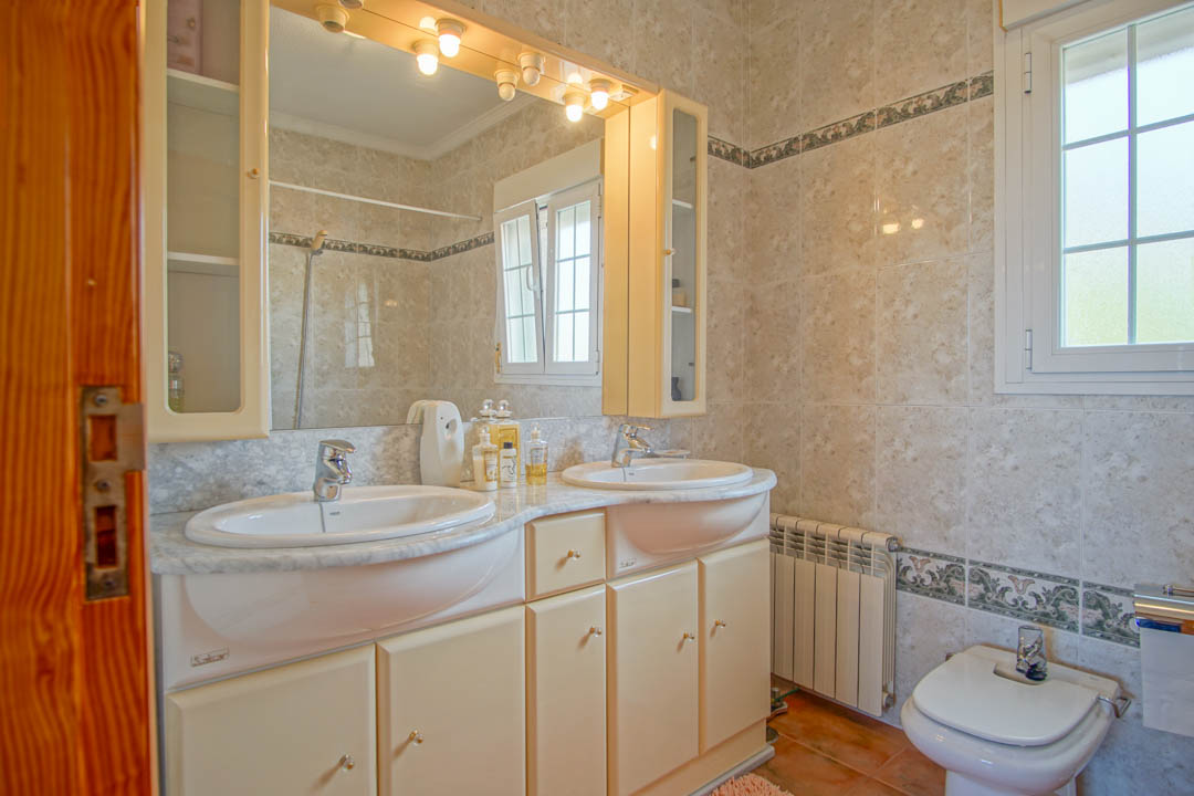 Baño con lavabo doble sobre encimera de mármol, muebles de madera maciza, espejo con iluminación integrada y paredes alicatadas con cenefa clásica.