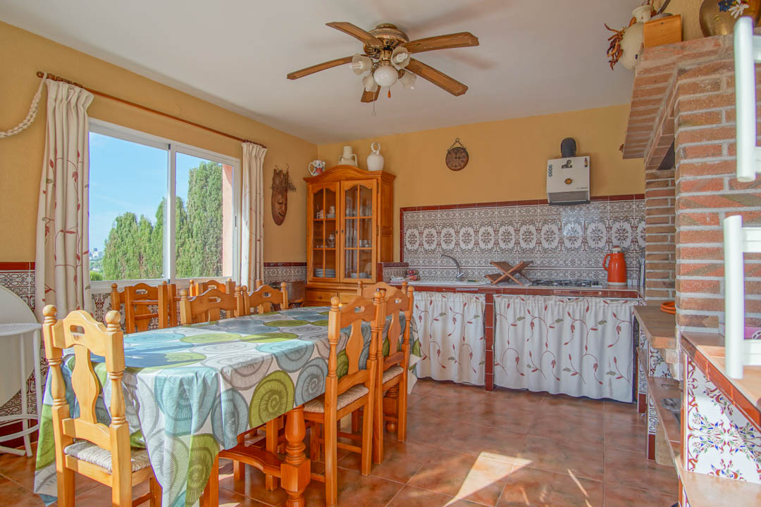 Cocina exterior cerrada con comedor de madera maciza, azulejos decorativos tradicionales y zona de barbacoa de obra integrada con vistas al jardín.