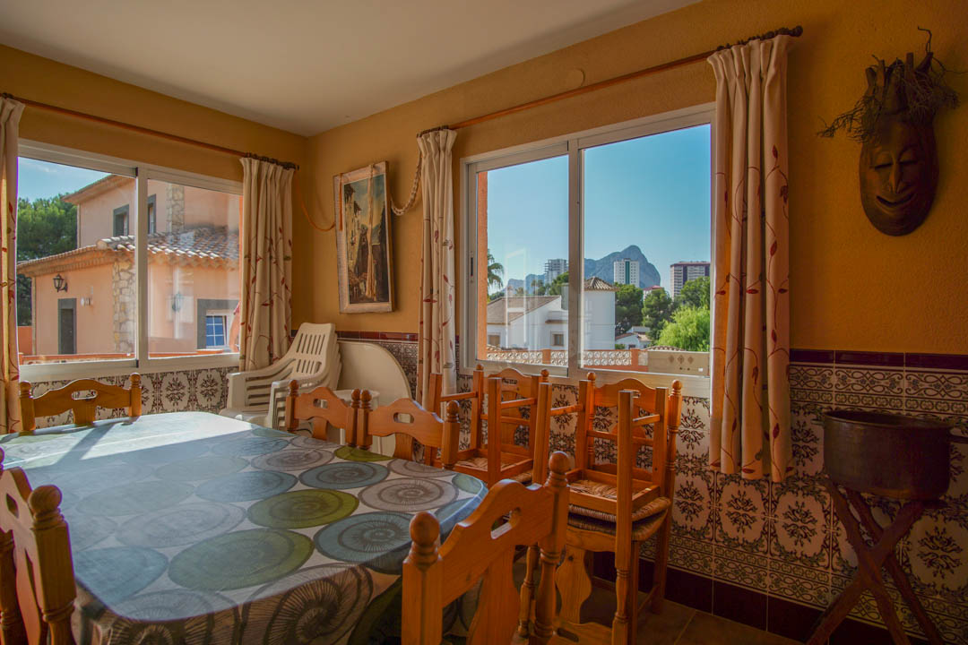 Comedor luminoso con mesa de madera maciza, zócalo de azulejos tradicionales y vistas despejadas al Peñón de Ifach a través de amplios ventanales.