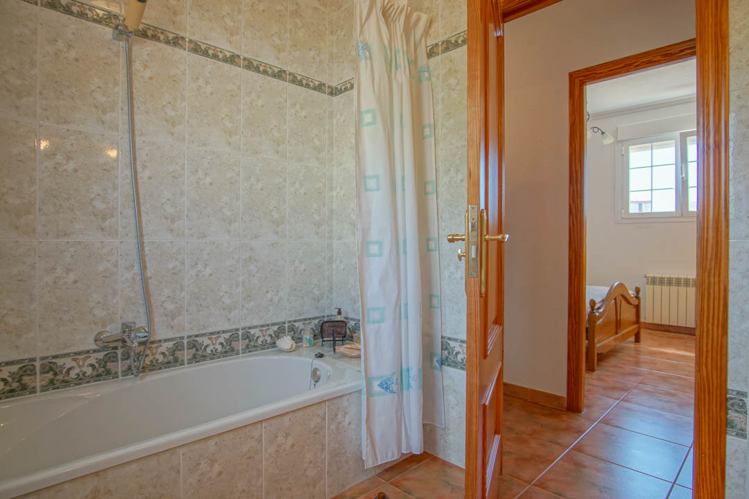 Cuarto de baño con bañera integrada, paredes alicatadas con cenefas decorativas y carpintería de madera maciza que conecta con un dormitorio luminoso.