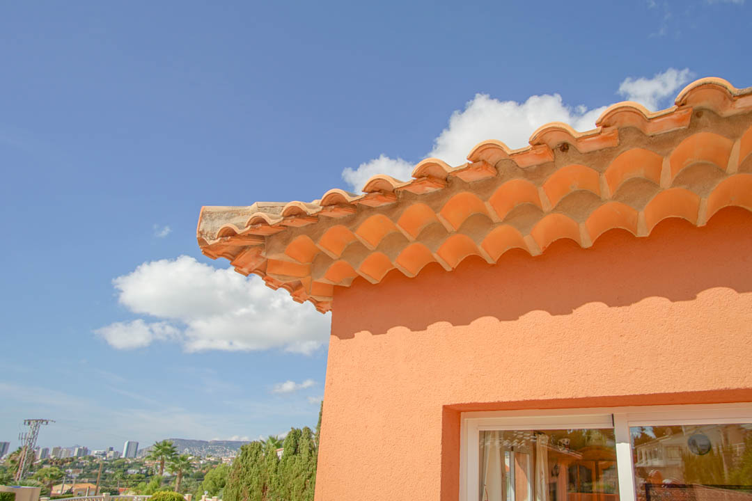 Detalle del tejado con tejas cerámicas y alero decorativo sobre la fachada terracota, con vistas despejadas al horizonte urbano y las montañas.