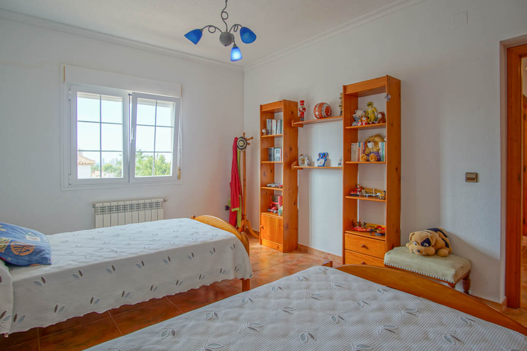 Dormitorio luminoso con dos camas individuales, estanterías de madera maciza y suelo de gres. Incluye radiador bajo la ventana y lámpara de techo decorativa.