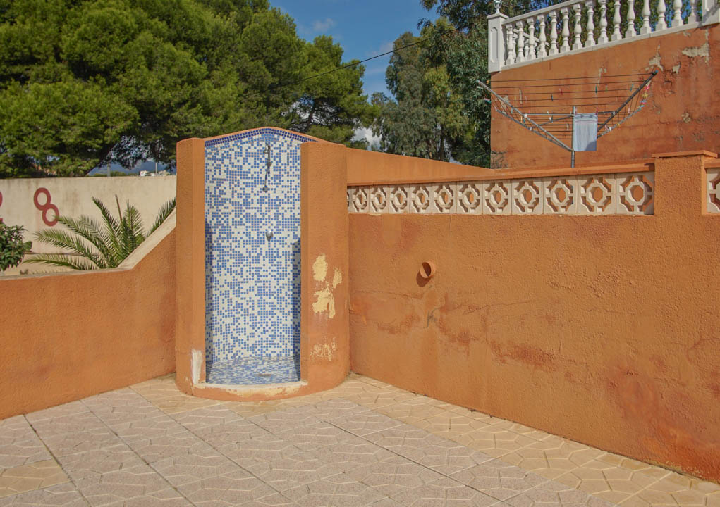 Ducha exterior con revestimiento de mosaico azul y blanco, situada en una zona de terraza pavimentada junto a muros de tono terracota con bloques decorativos.
