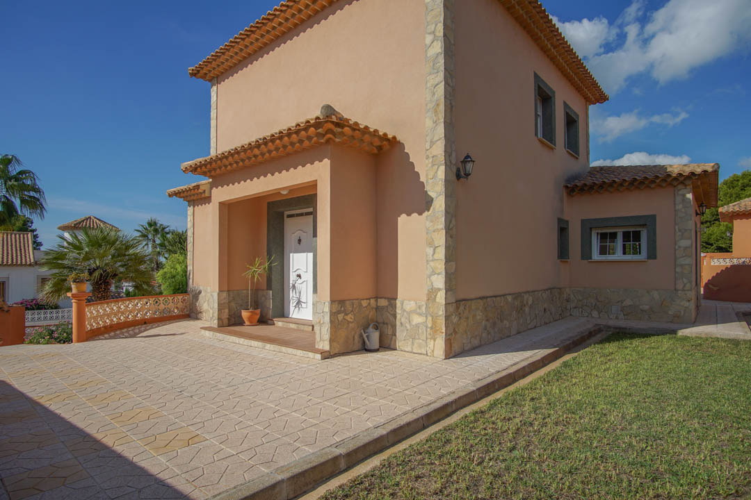 Fachada exterior con zócalo de piedra natural, entrada principal bajo porche y pavimento cerámico decorativo junto a zona de jardín.