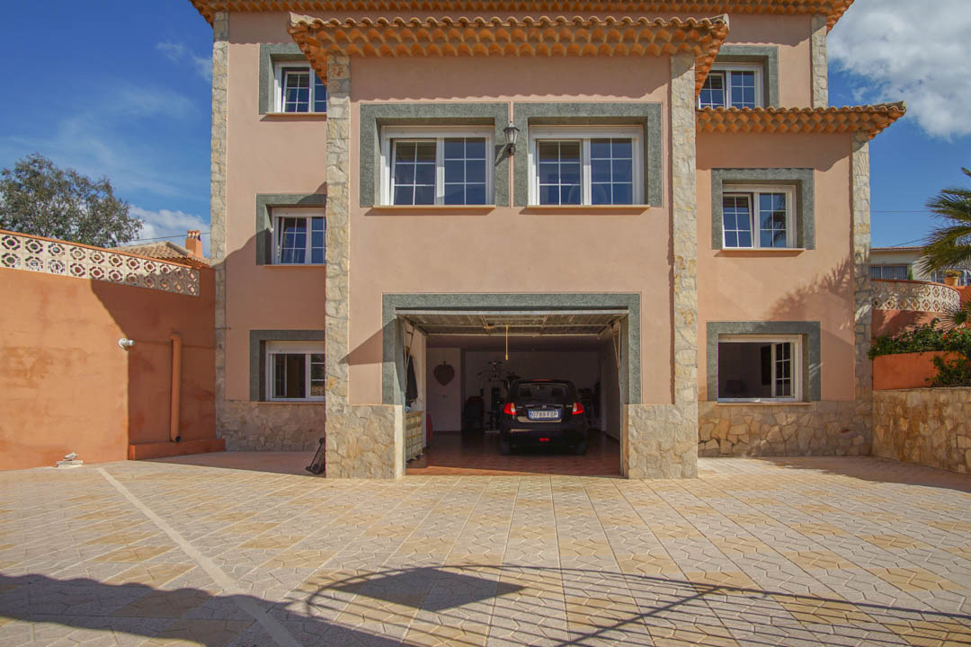 Fachada frontal de la villa con amplio aparcamiento pavimentado y acceso al garaje integrado. Destacan los acabados en piedra natural y la sólida estructura de tres niveles.