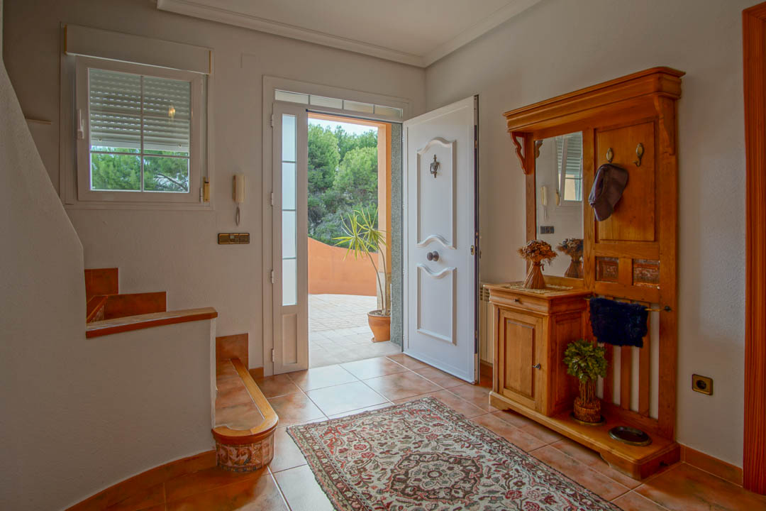 Hall de entrada con puerta principal blanca, suelos de gres rústico y un mueble recibidor de madera maciza con espejo. La luz natural ilumina el espacio desde el acceso exterior.