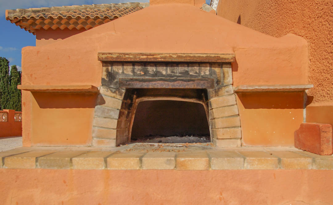 Horno de leña exterior integrado en estructura de obra con acabado terracota y embocadura de ladrillo refractario bajo tejado de teja tradicional.