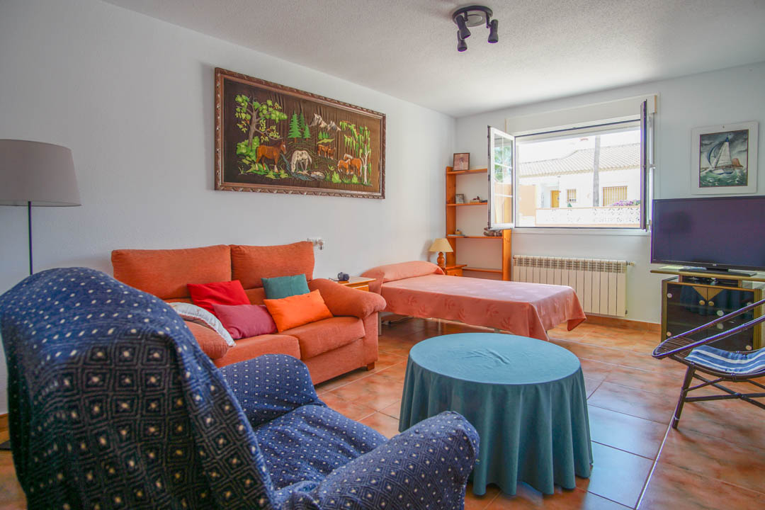 Sala de estar funcional con suelos cerámicos, zona de descanso con sofá y cama individual, iluminada por un gran ventanal con vistas despejadas.