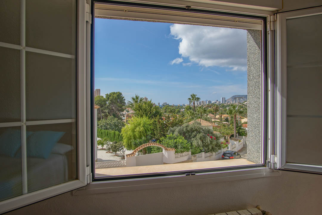 Vista despejada desde el dormitorio hacia el entorno residencial y el horizonte costero a través de una ventana de carpintería blanca.