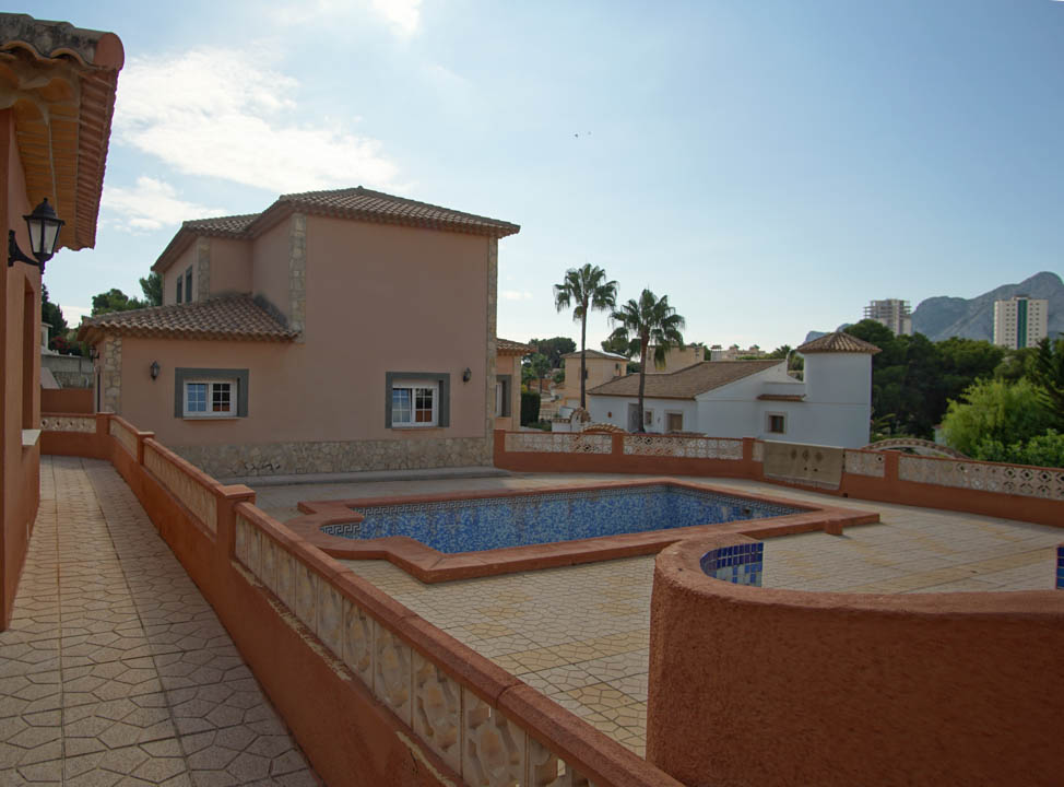 Vista exterior de la piscina con azulejos de mosaico azul, rodeada por una amplia terraza pavimentada y muros de color terracota con detalles en piedra natural.