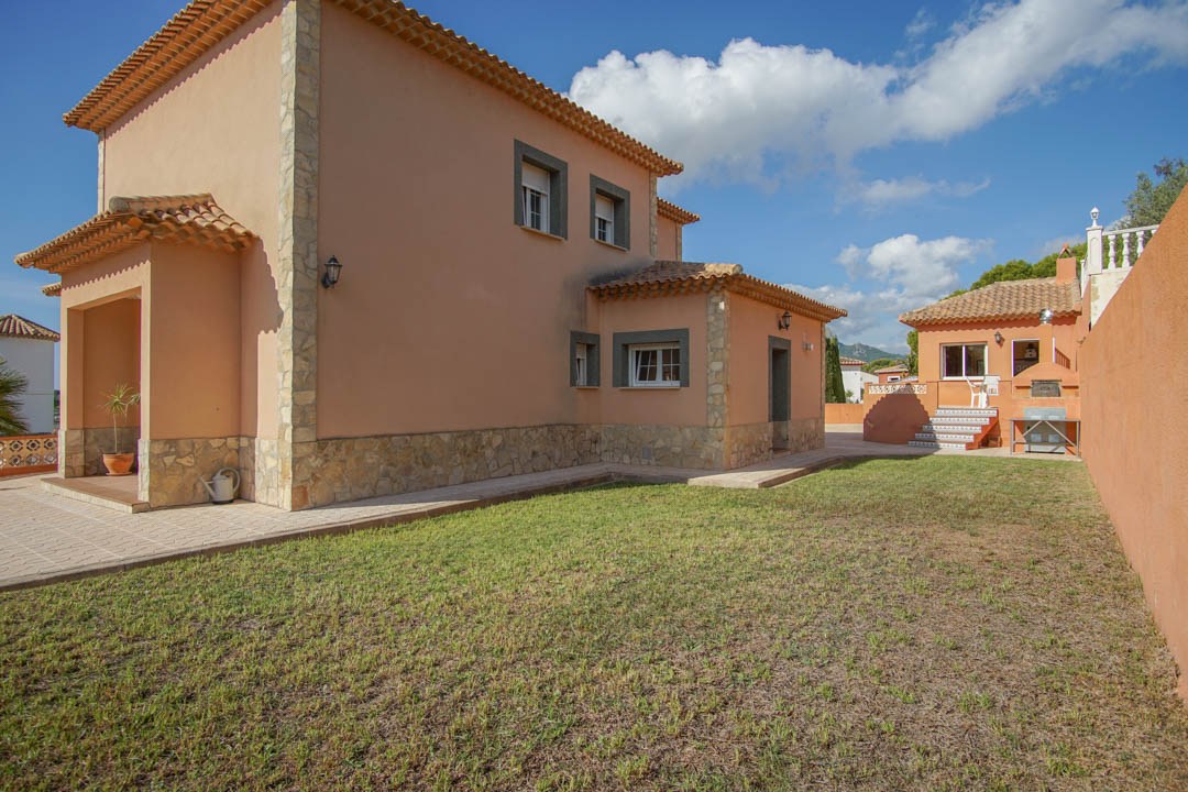 Vista exterior de la villa con fachada en tonos tierra, zócalo de piedra natural y jardín. Al fondo destaca la cocina exterior independiente con horno de leña.