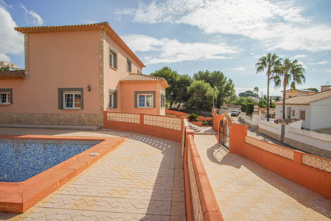 Vista exterior de la villa mostrando la fachada en tonos tierra con detalles de piedra, la zona de la piscina y el acceso pavimentado desde la calle.