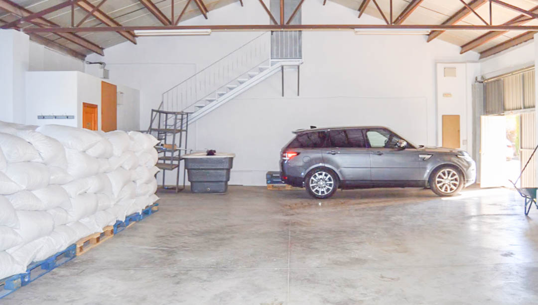 Amplio almacén industrial con suelos de hormigón pulido, vigas metálicas a la vista y espacio para vehículos y almacenamiento de productos agrícolas sobre palés.