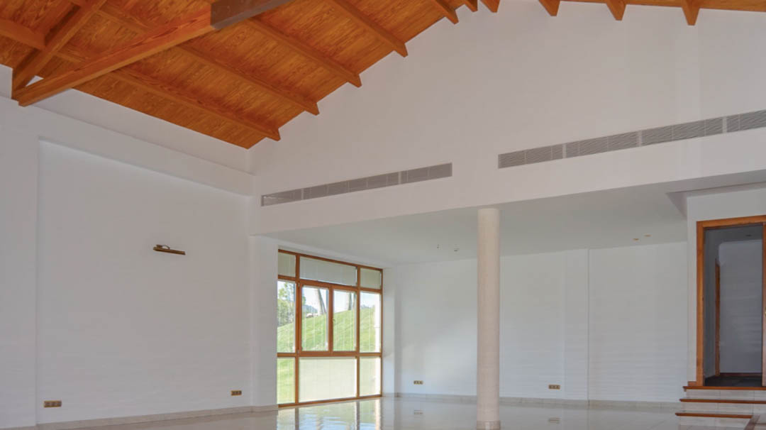 Amplio salón con techos altos de madera vista, suelos de gres pulido y grandes ventanales. Incluye climatización integrada y una columna estructural elegante.
