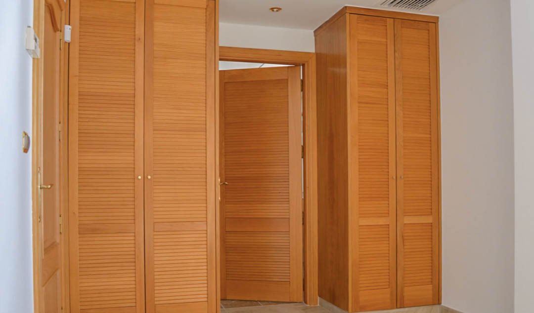 Armarios empotrados de madera de alta calidad con puertas de lamas tipo mallorquina, integrados en un pasillo con carpintería a juego y suelos cerámicos.